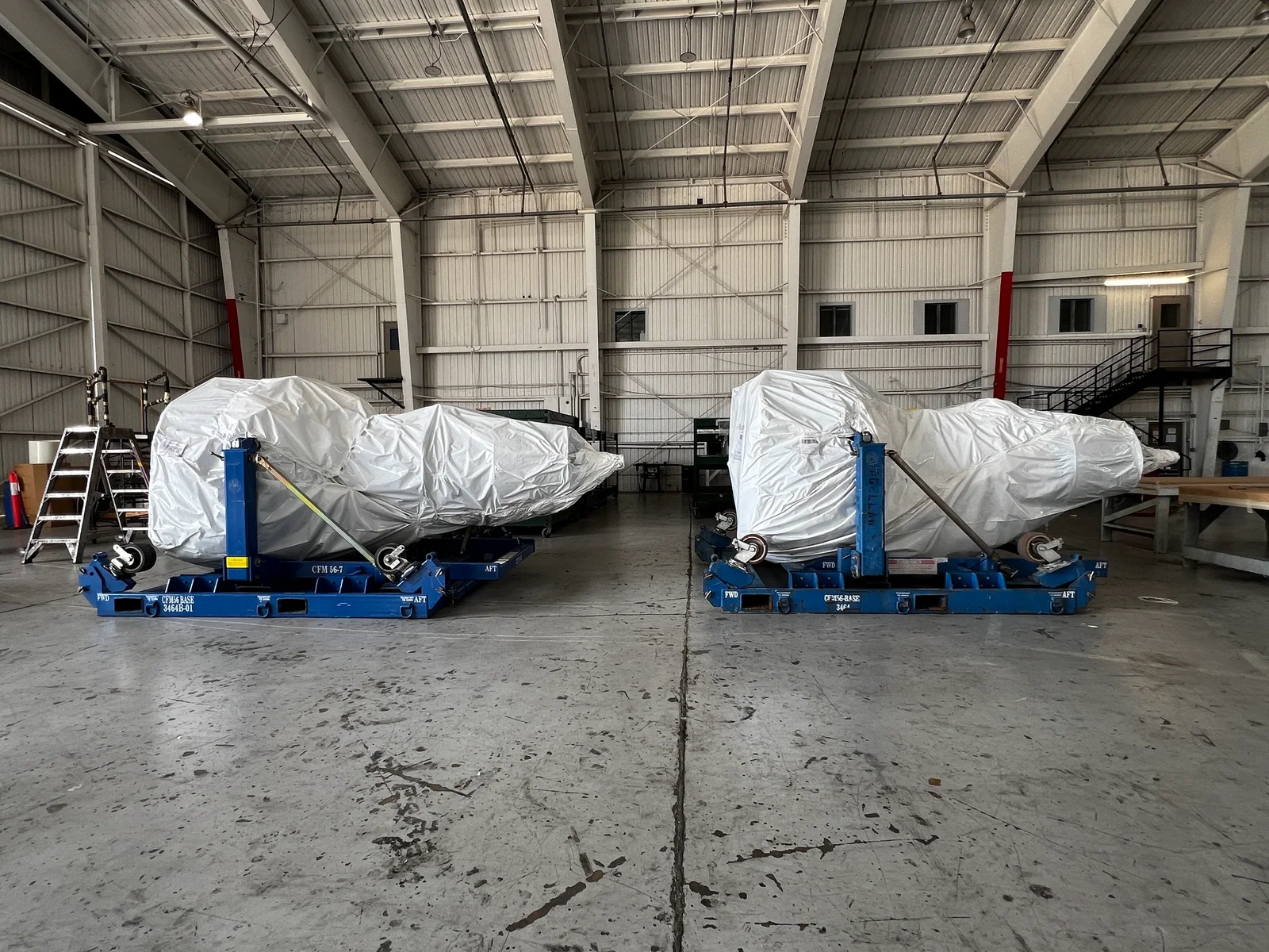 Two large wrapped aerospace components on blue transport bases inside a warehouse.