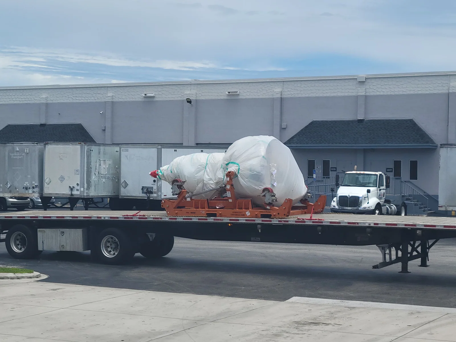 Large wrapped component secured on a long flatbed trailer outdoors.