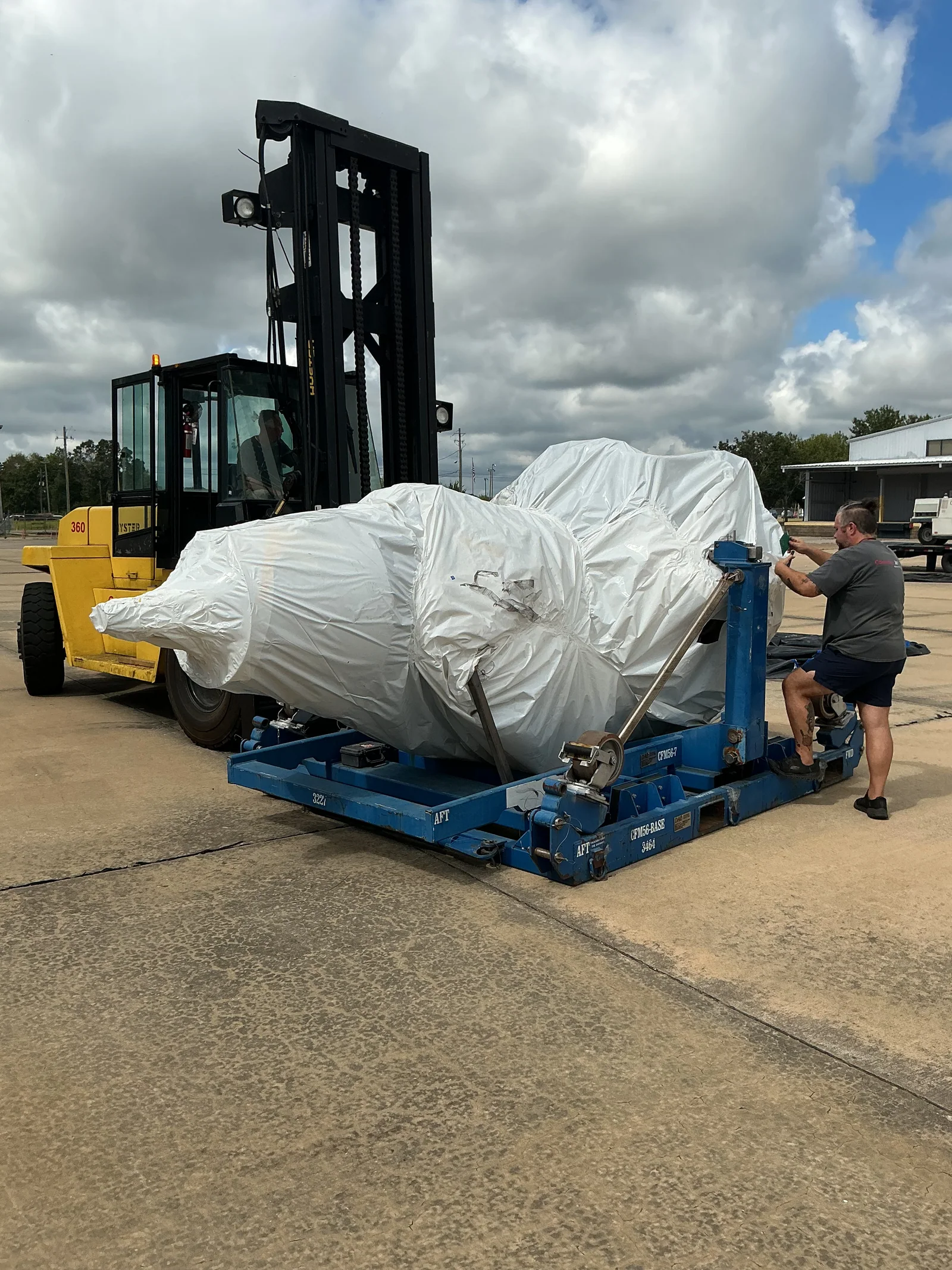 Forklift and worker beside a large white wrapped component on a blue support frame.