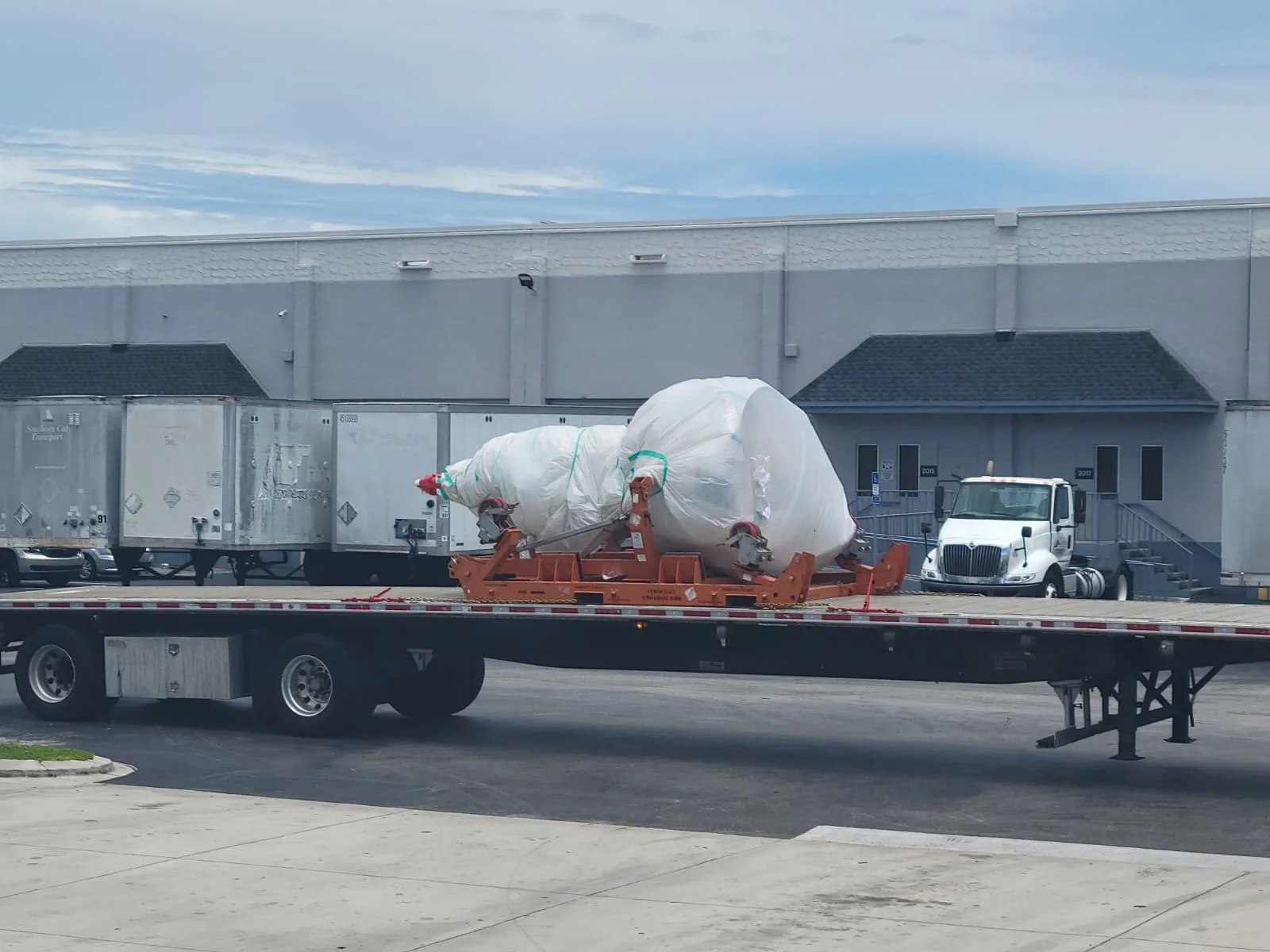 Wrapped component on a road trailer beside an industrial building.