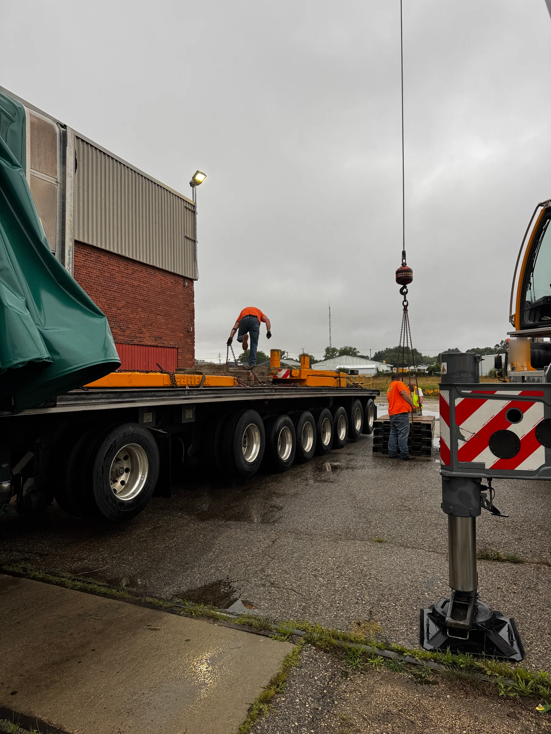 Workers beside a long trailer and a crane hook in an industrial yard.