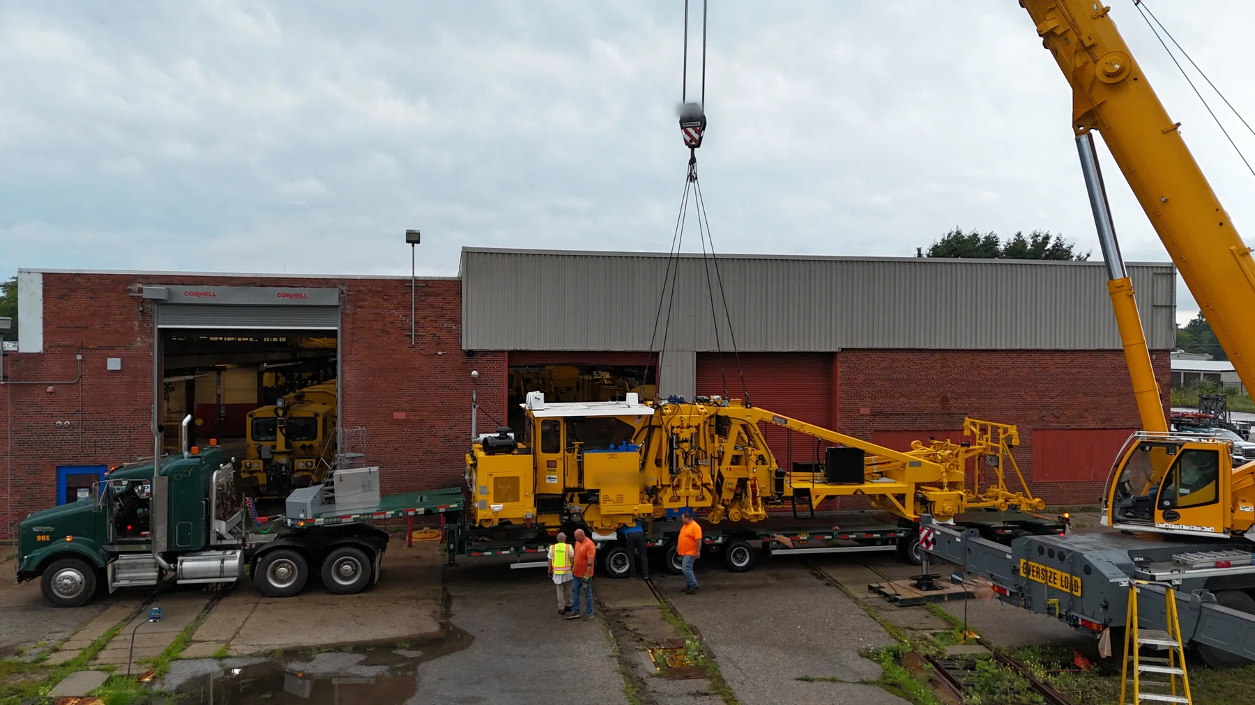 Wide view of a crane beside railway equipment on a trailer with workers nearby.