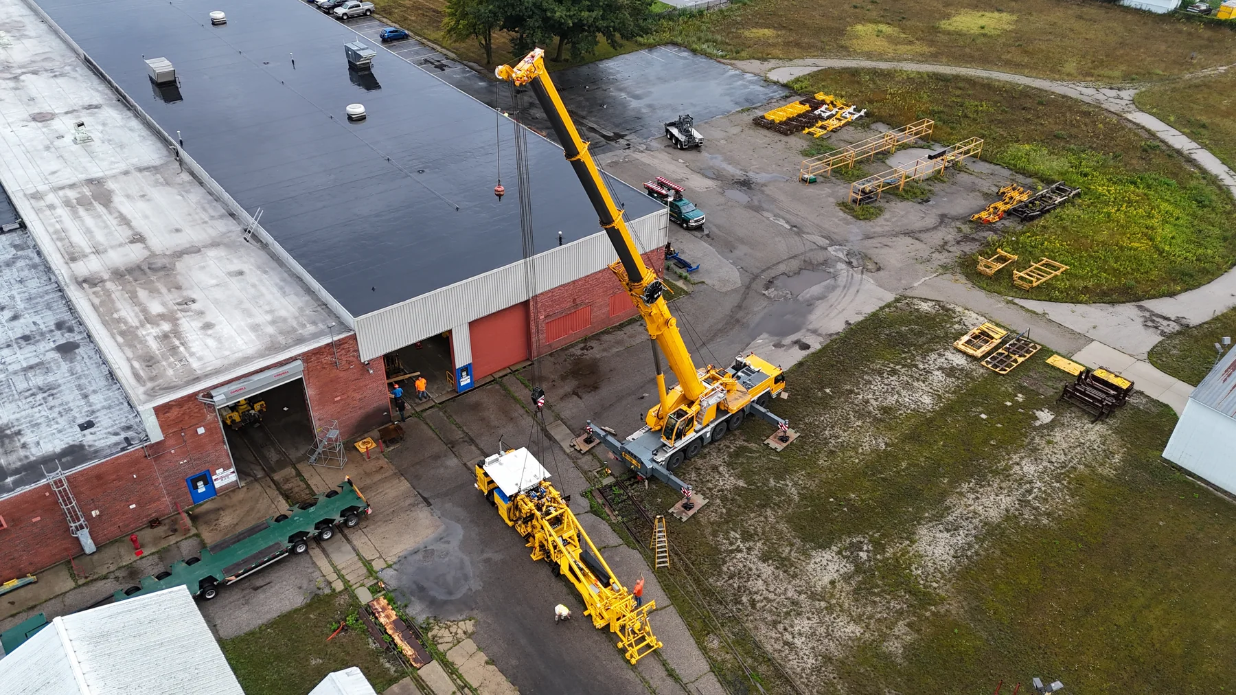 Aerial view of a crane lifting yellow railway equipment beside a long road trailer.